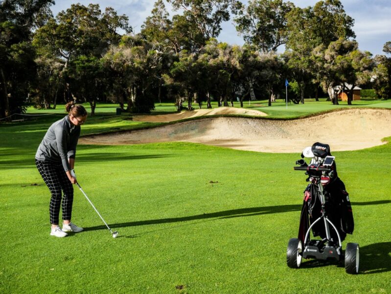 Female golfer aiming at ball in a golf course surrounded by a bunker and a buggy cart